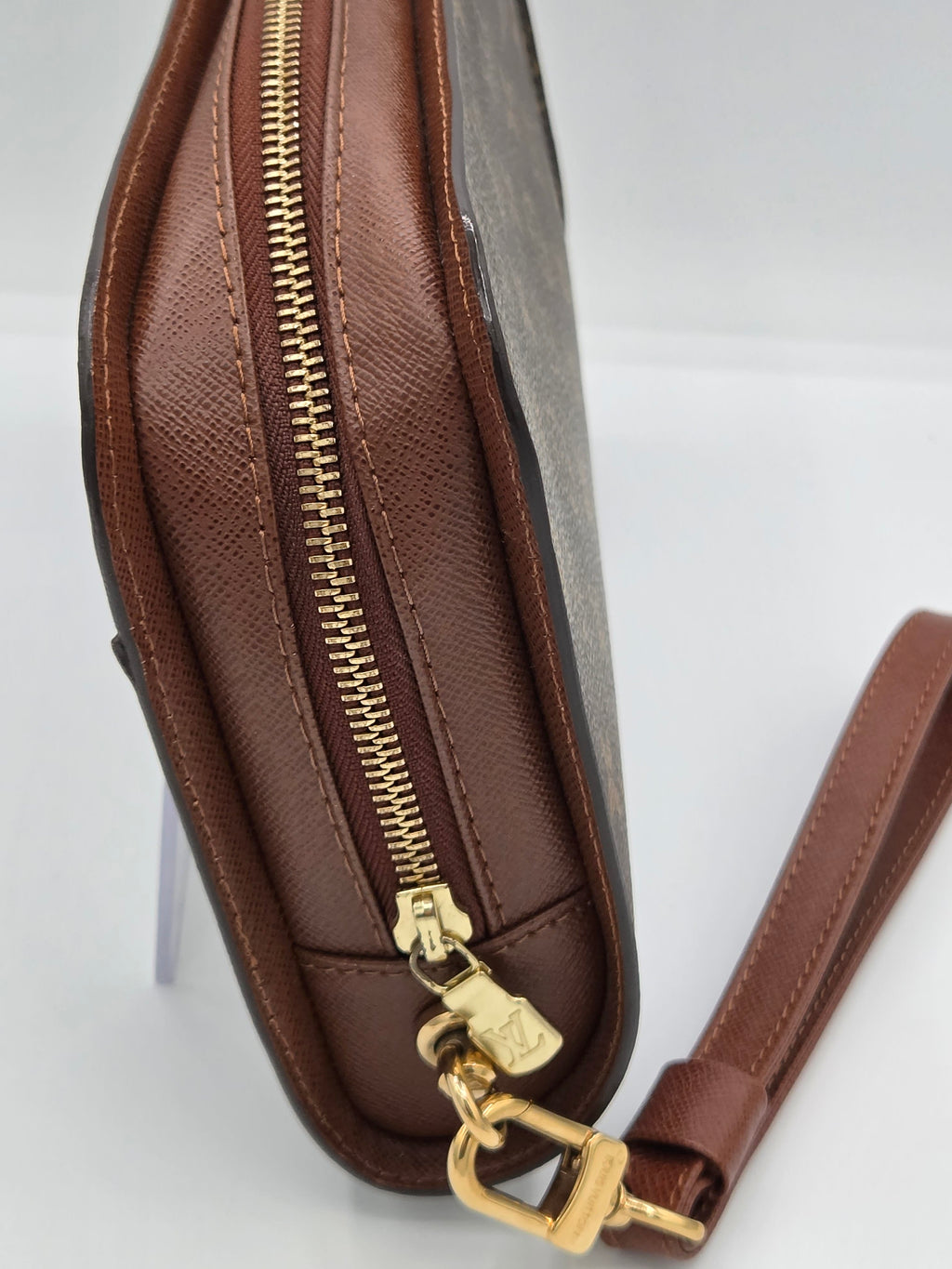 A close-up of a brown leather clutch or wristlet bag with a gold zipper, gold hardware, and a matching brown leather wrist strap on a white background.