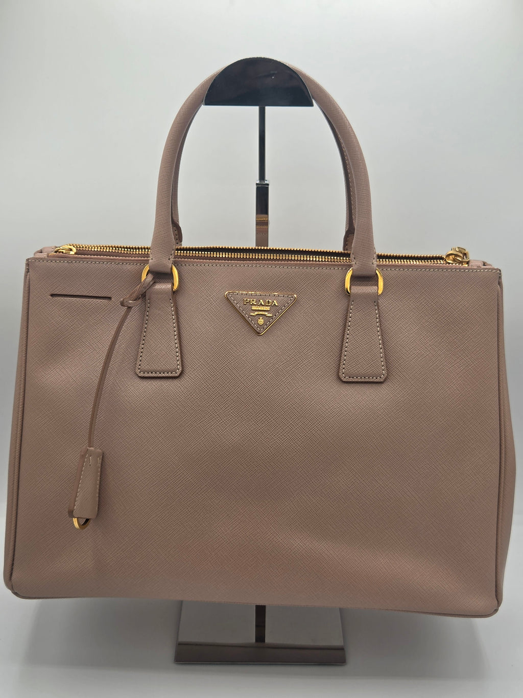A Prada taupe leather handbag with gold-tone hardware, displayed on a black stand against a light gray background.