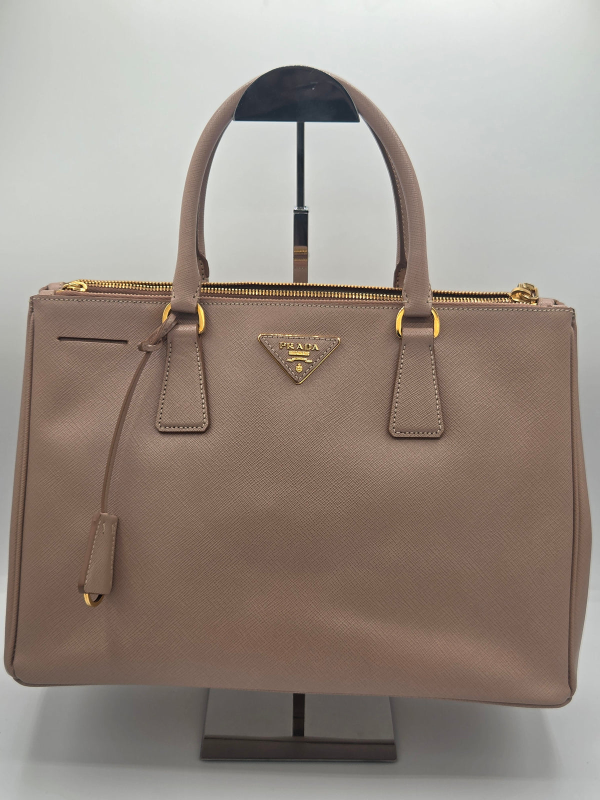A Prada taupe leather handbag with gold-tone hardware, displayed on a black stand against a light gray background.