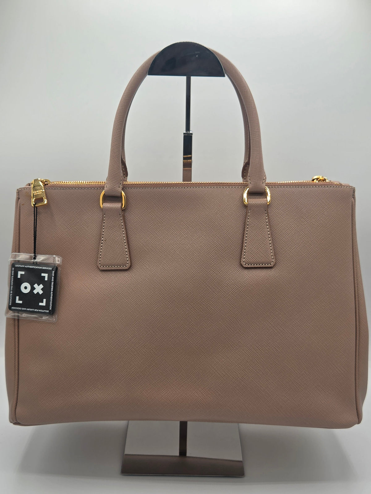 A taupe brown textured leather handbag with gold-tone hardware, displayed on a black stand against a light gray background.