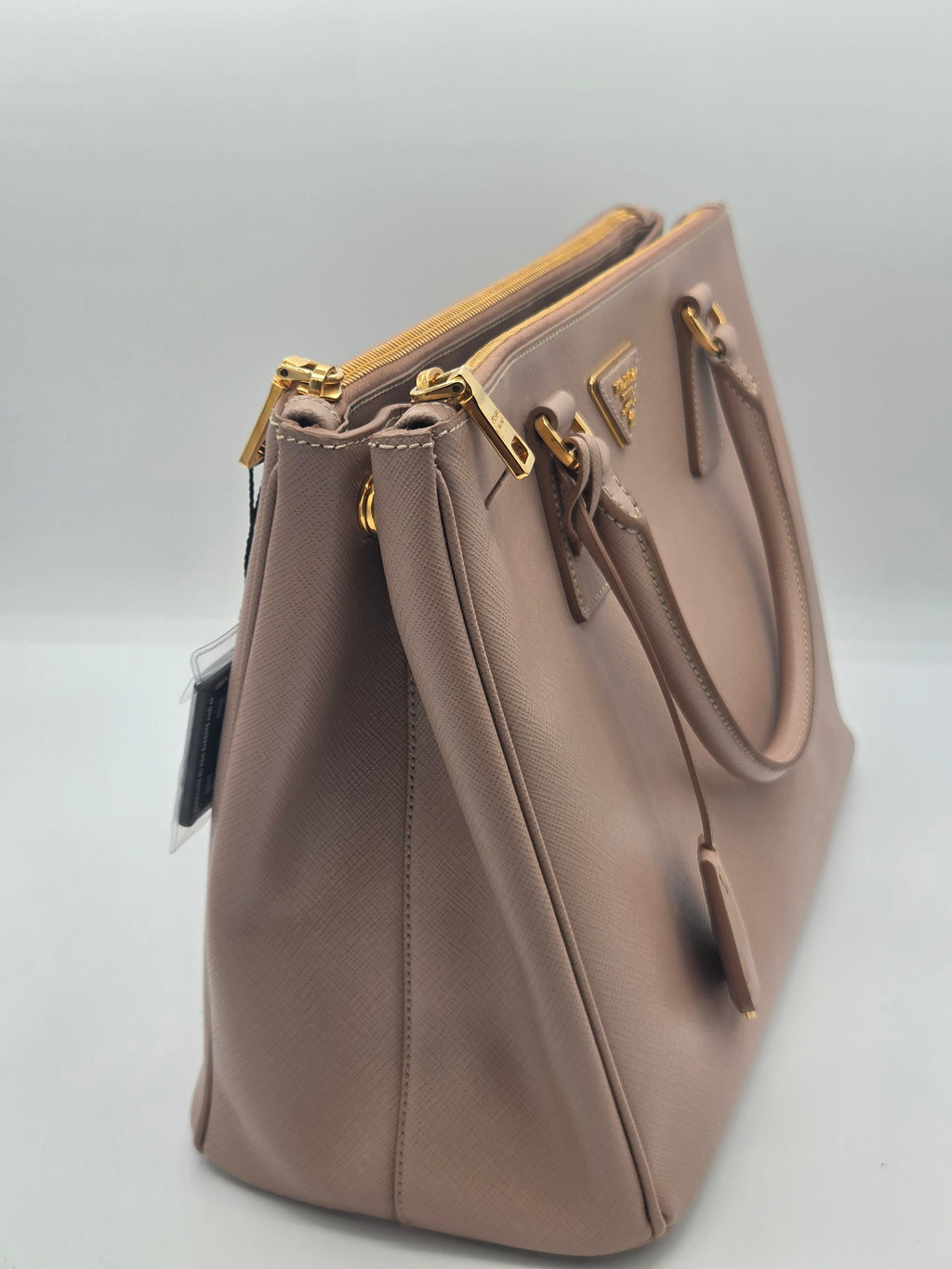 A Prada shoulder bag in taupe pebbled leather with gold-tone hardware and a triangular Prada logo plaque on the front, shown on a light gray background.