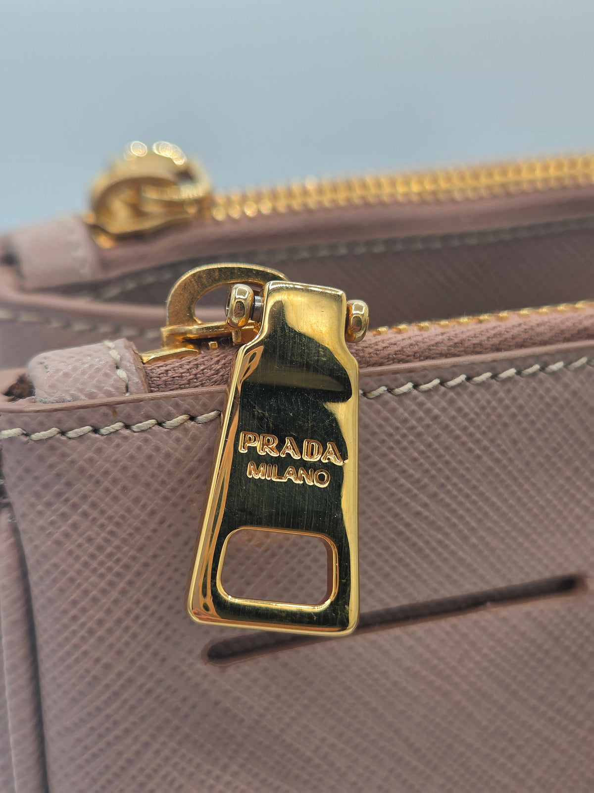 Close-up of a Prada Milano handbag in taupe leather with a polished gold-tone zipper pull engraved with 'PRADA MILANO'.