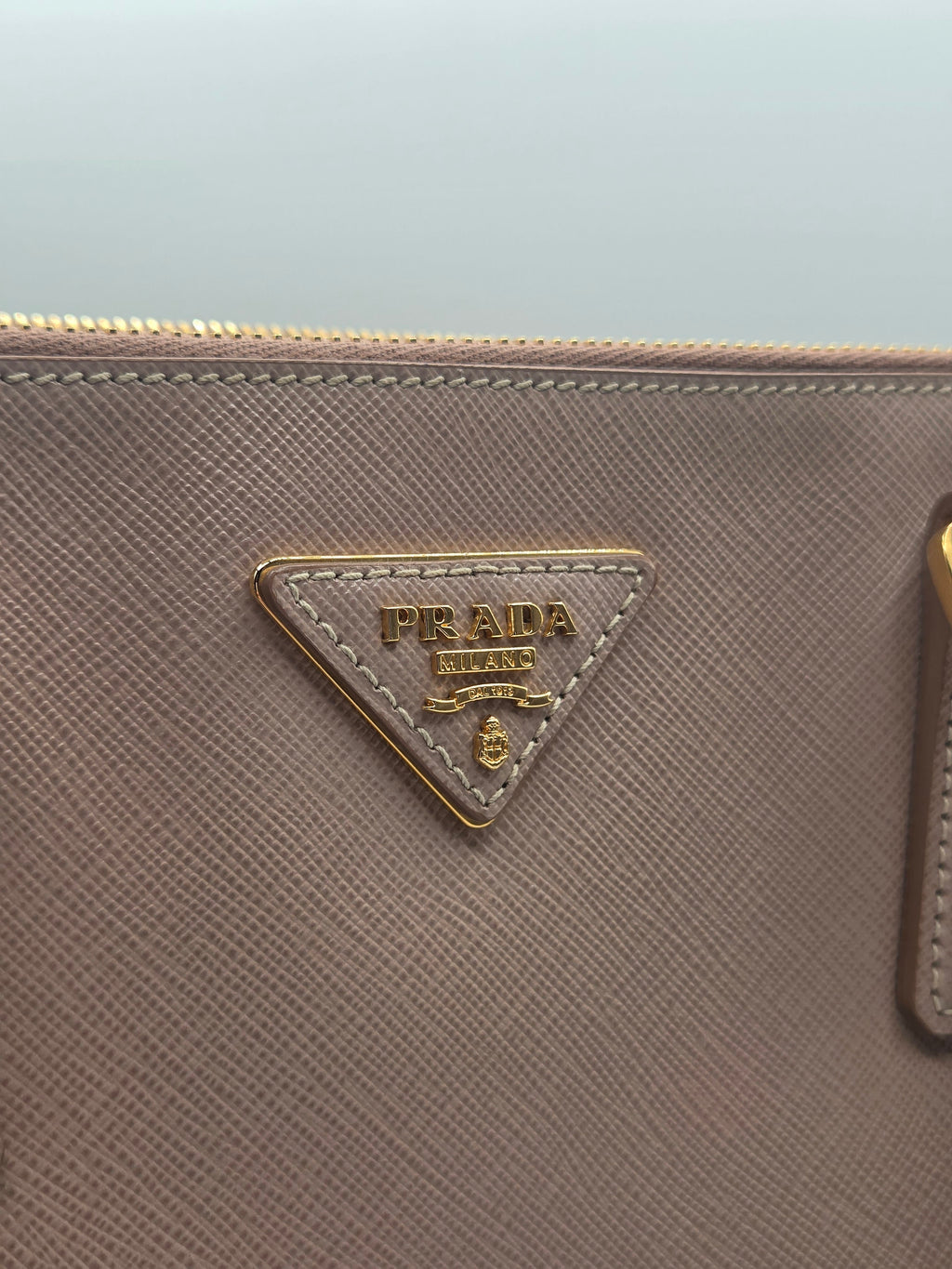 Close-up of a Prada pouch in textured taupe leather with the iconic gold-tone triangular logo plaque on the front.
