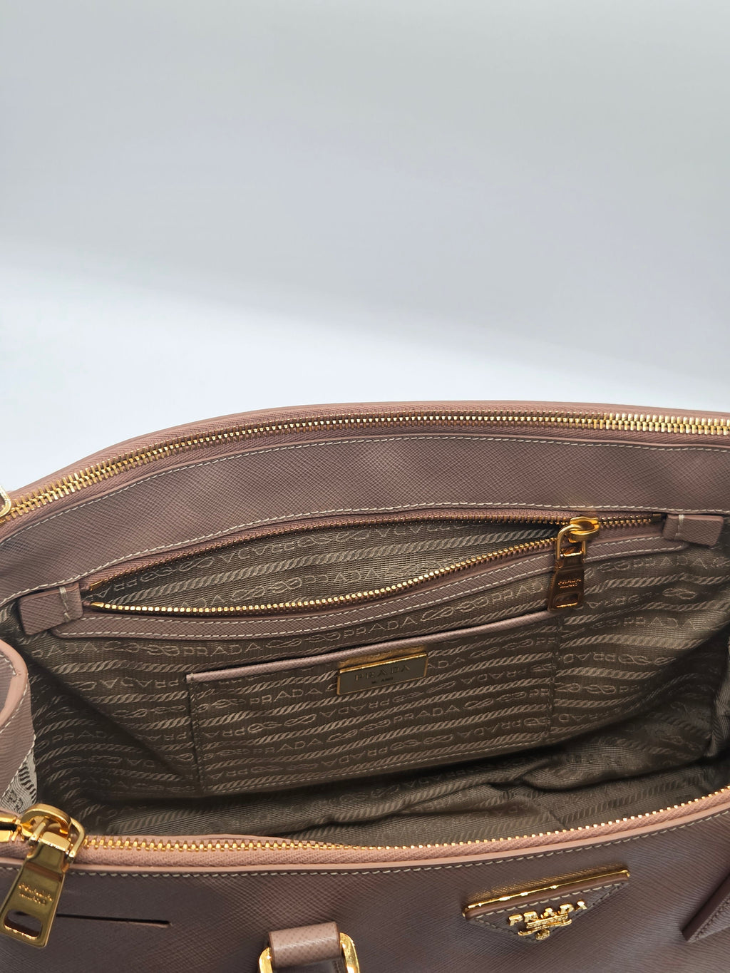 Close-up of the interior of a Prada handbag in taupe-brown coated canvas with gold-tone hardware, showing the branded monogrammed lining and zip compartments.