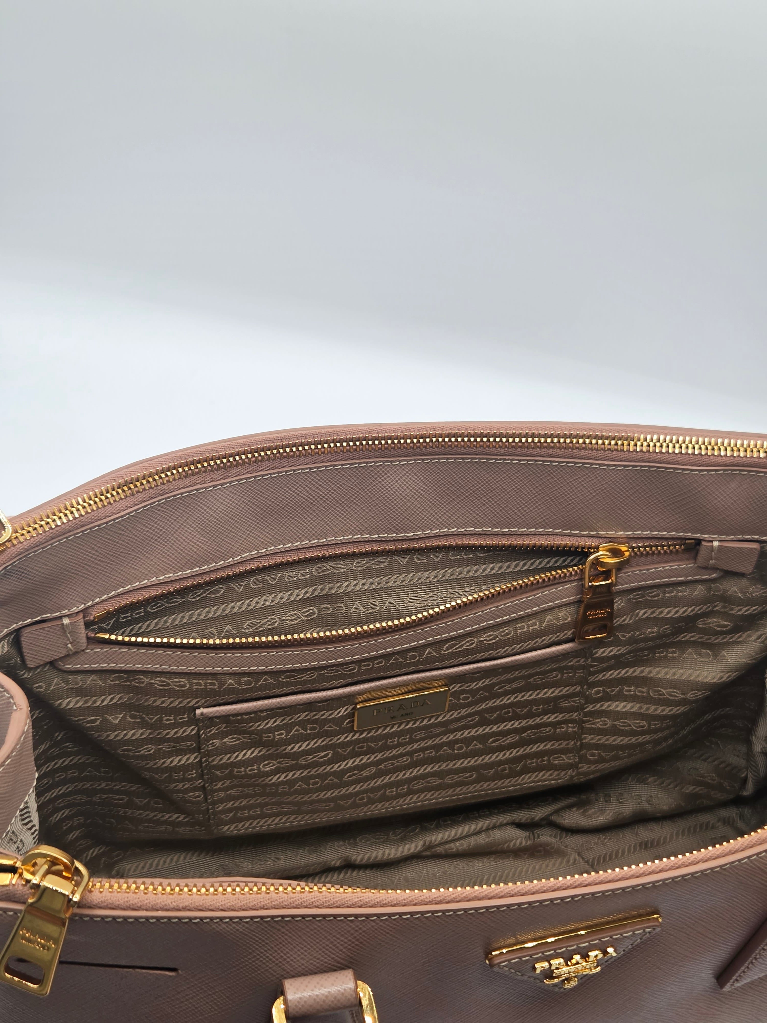Close-up of the interior of a Prada handbag in taupe-brown coated canvas with gold-tone hardware, showing the branded monogrammed lining and zip compartments.