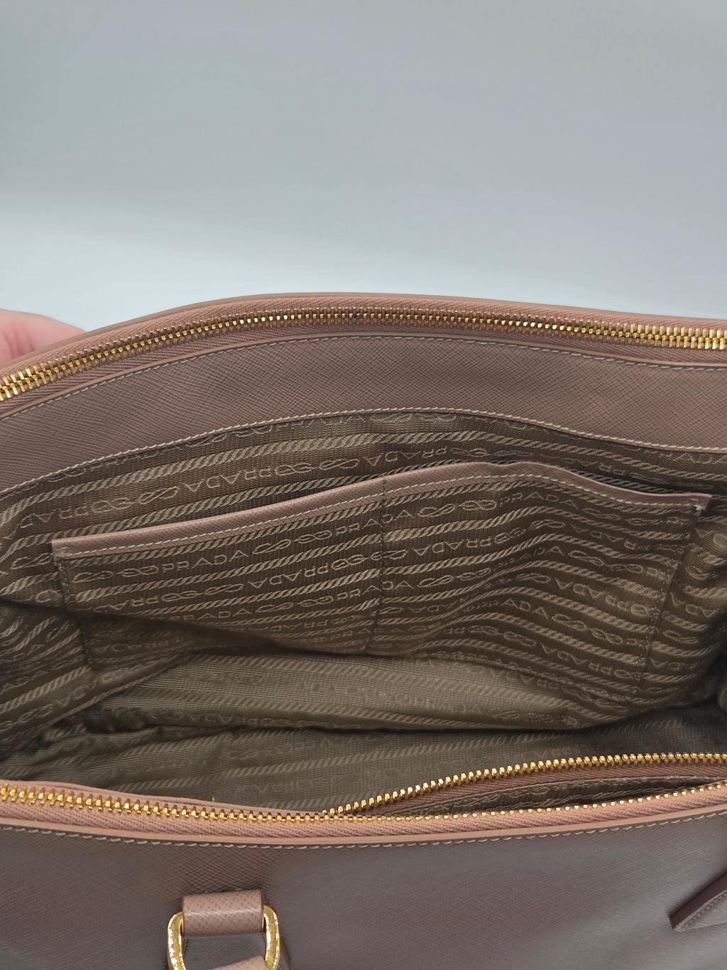 Close-up of the interior of a Prada handbag, showing a brown and beige jacquard patterned lining with gold-tone zippers and a small gold hardware detail.