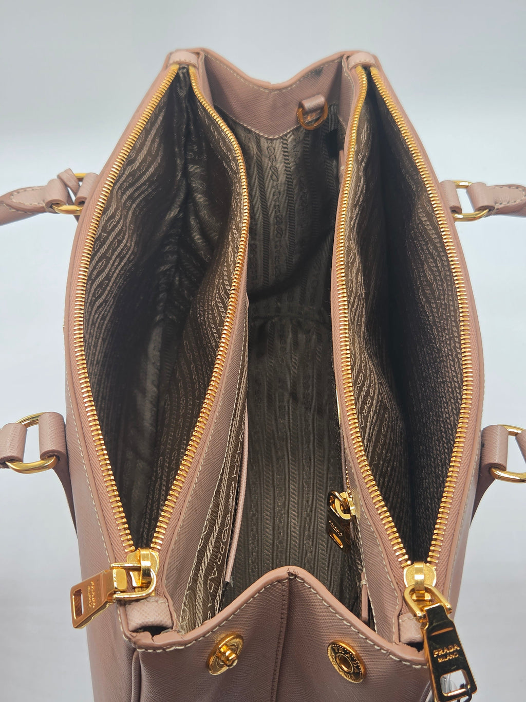 An open taupe leather handbag with gold zippers and hardware, showing a dark patterned interior lining.
