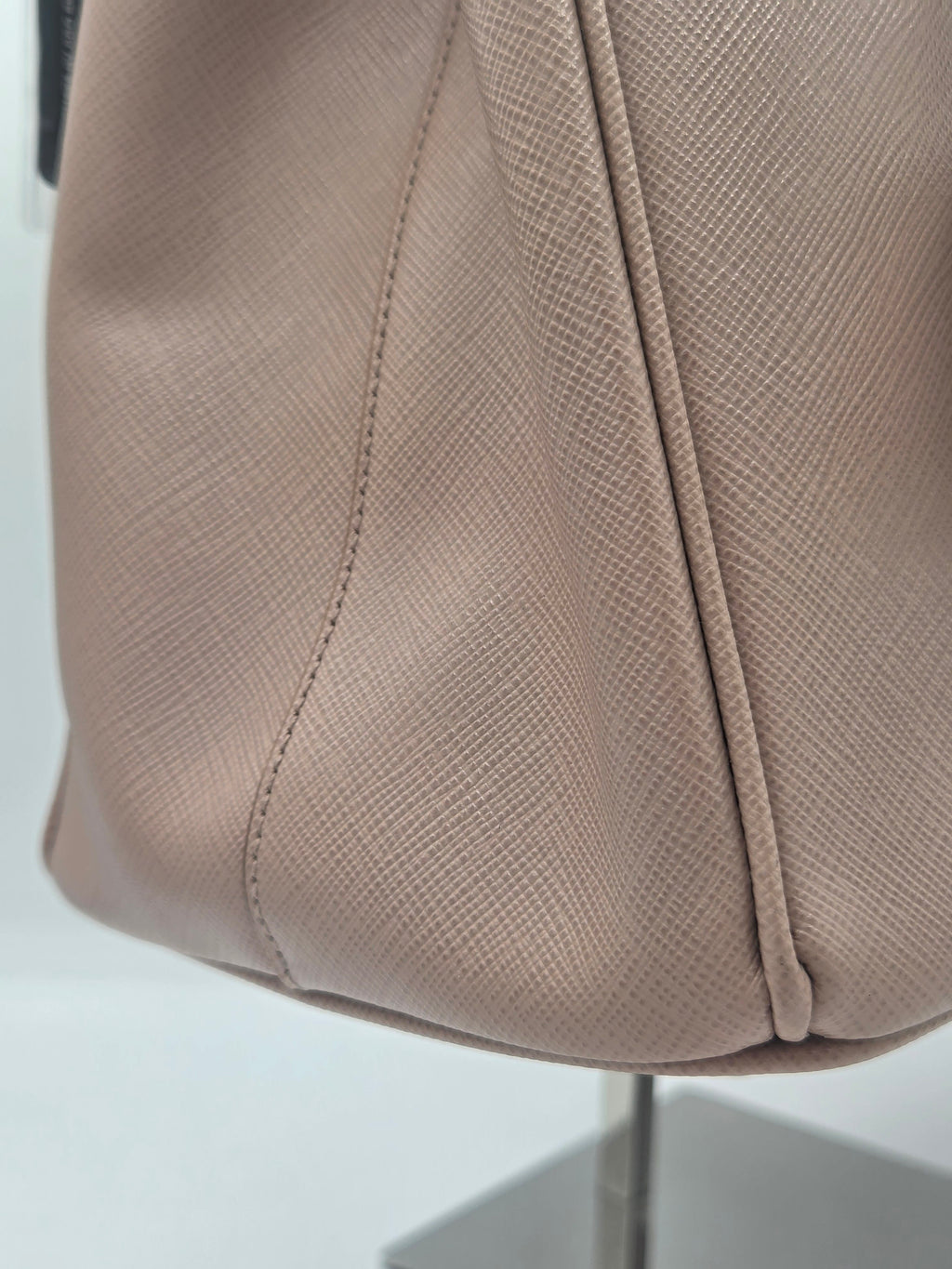 Close-up of a taupe textured leather bag corner with visible stitching and a metallic stand base against a light gray background.