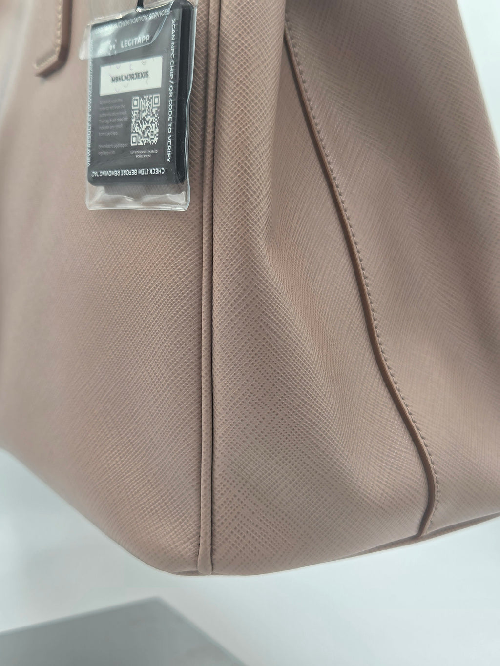 Close-up of a taupe-brown textured leather handbag corner with a hangtag attached, showing stitched seam and material texture against a light neutral background.