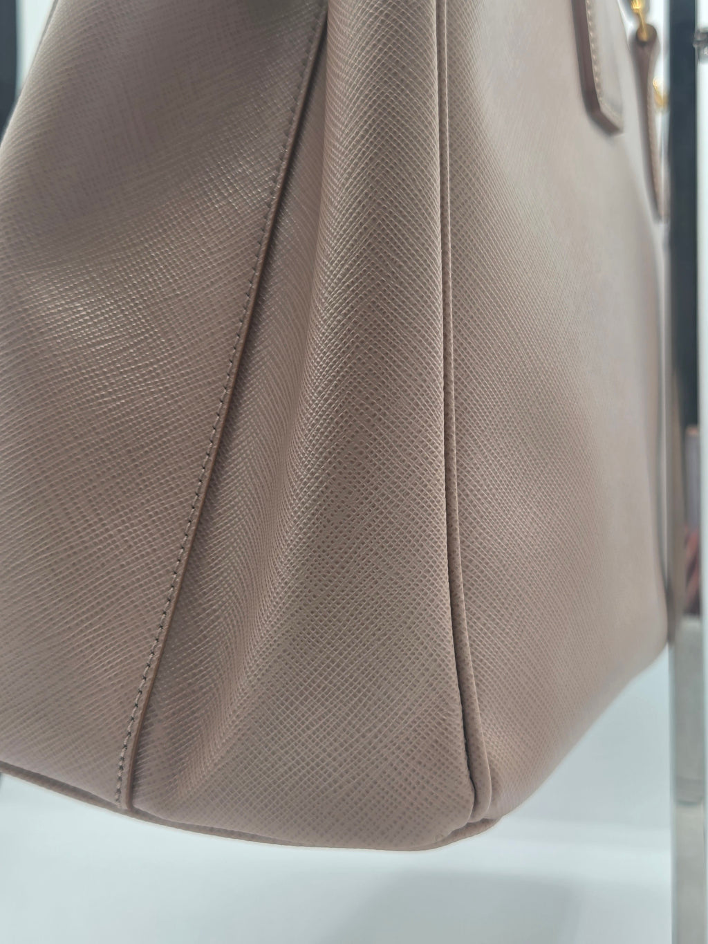 Close-up of a taupe textured leather handbag corner showing the side panel, stitching, and a small gold-tone hardware detail.