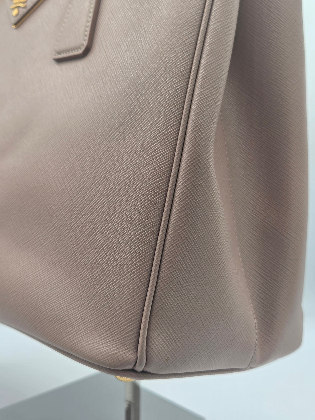 Close-up of a taupe-brown textured leather handbag corner with gold-tone hardware and clean stitching, displayed on a stand against a light neutral background.