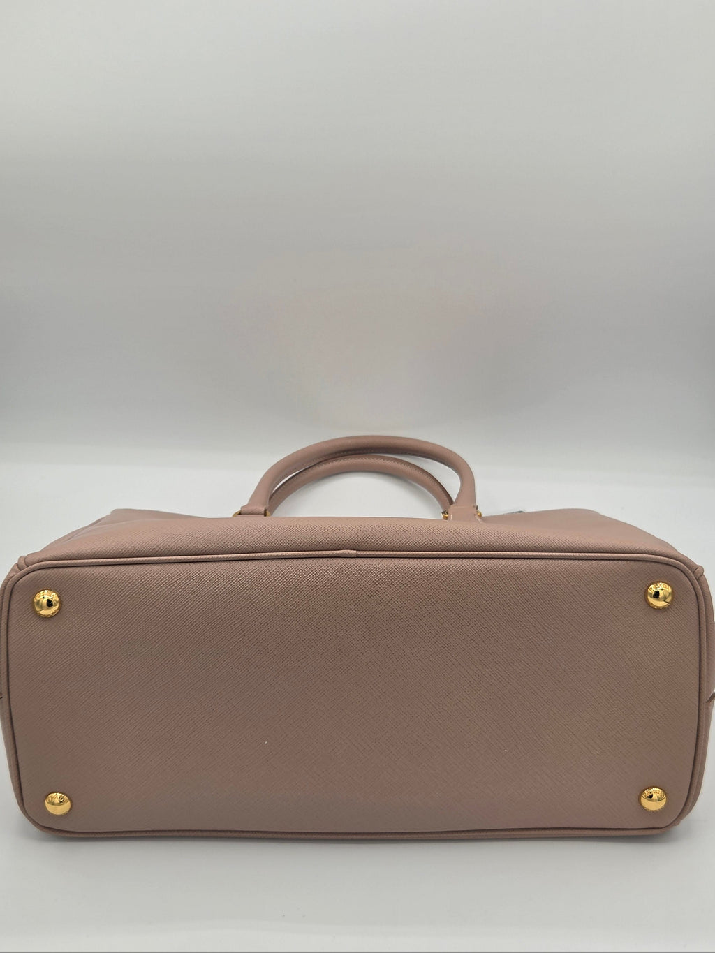 A structured top-handle handbag in dusty blush pink textured leather, shown from the back to display the gold-tone metal studs and base.