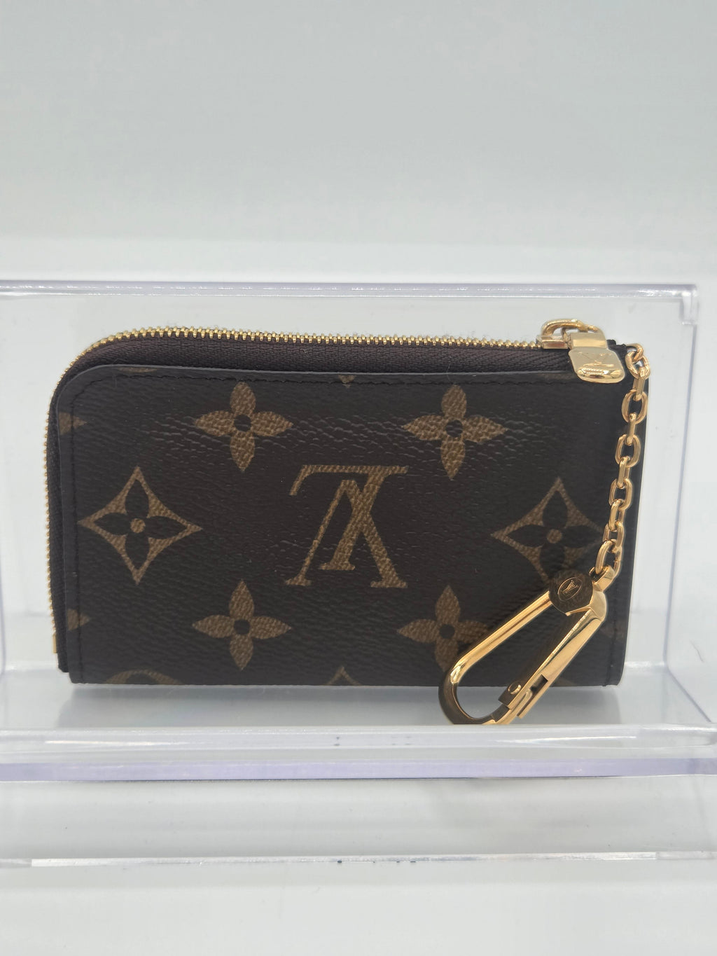 A Louis Vuitton monogram canvas zip-around wallet with a gold-tone chain and carabiner clip, displayed on a clear acrylic stand.