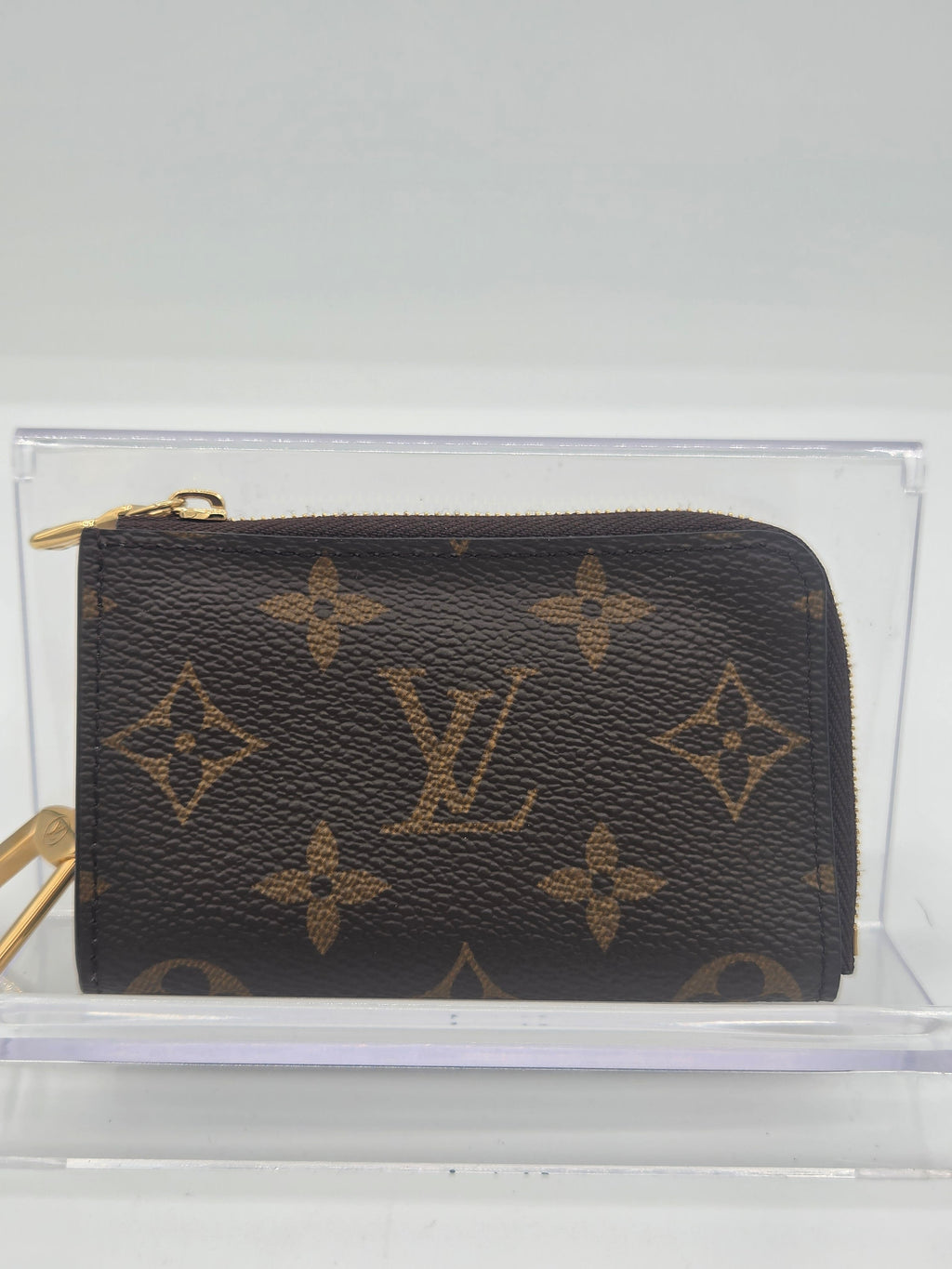 A Louis Vuitton monogram zip-around coin pouch in dark brown coated canvas with tan monogram motifs and gold-tone hardware, displayed inside a clear acrylic case.