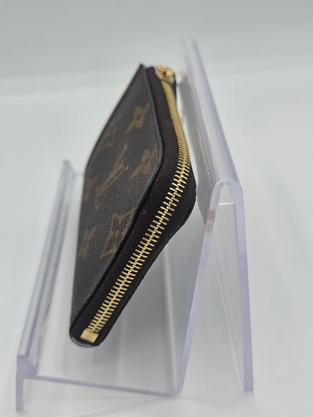 A Louis Vuitton monogram canvas zip-around pouch with gold-tone hardware, displayed on a clear acrylic stand against a white background.