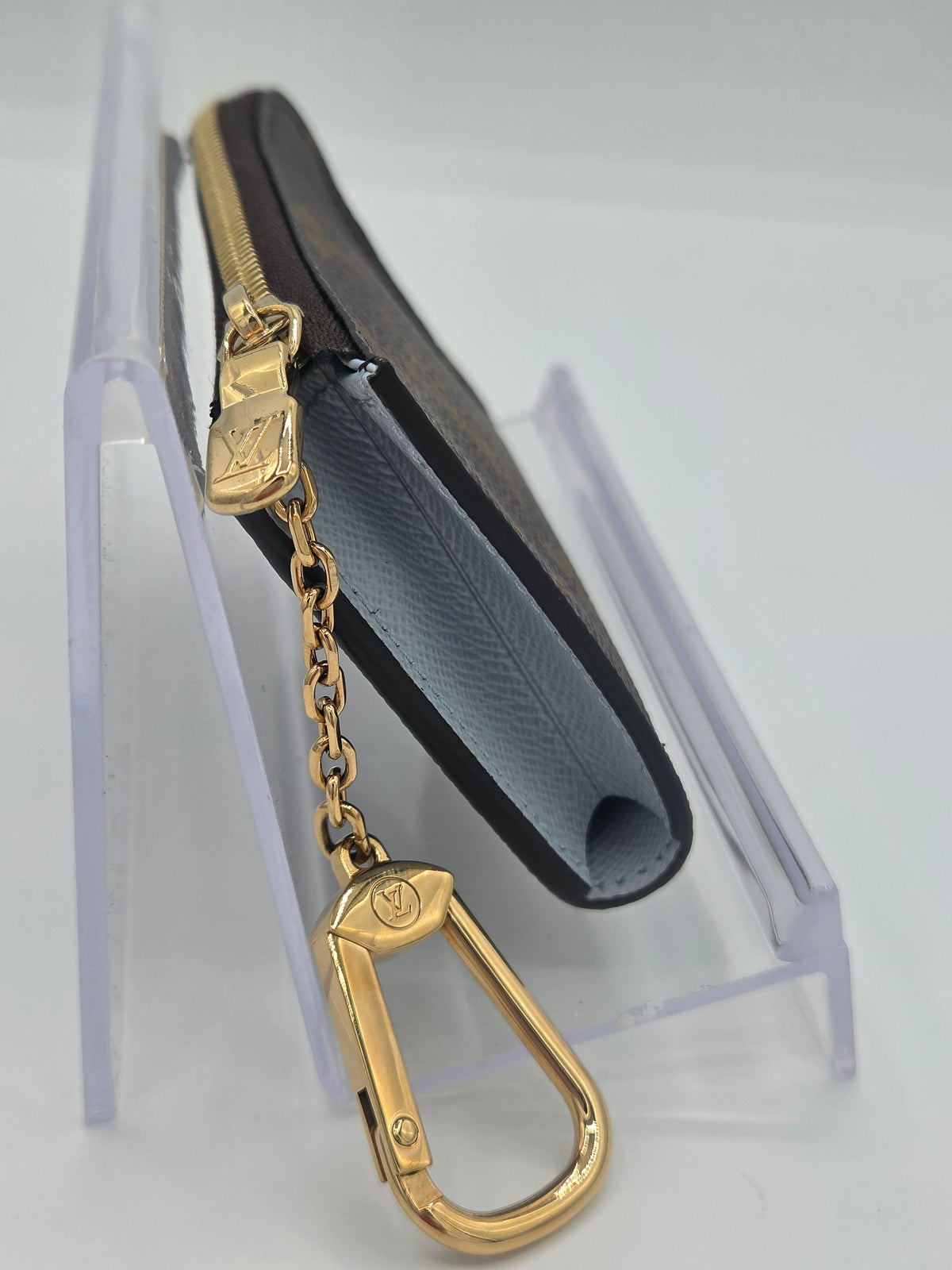 A Louis Vuitton monogram card holder wallet in dark brown and light blue-gray canvas, featuring a gold-tone chain and carabiner keychain, displayed on a clear acrylic stand.