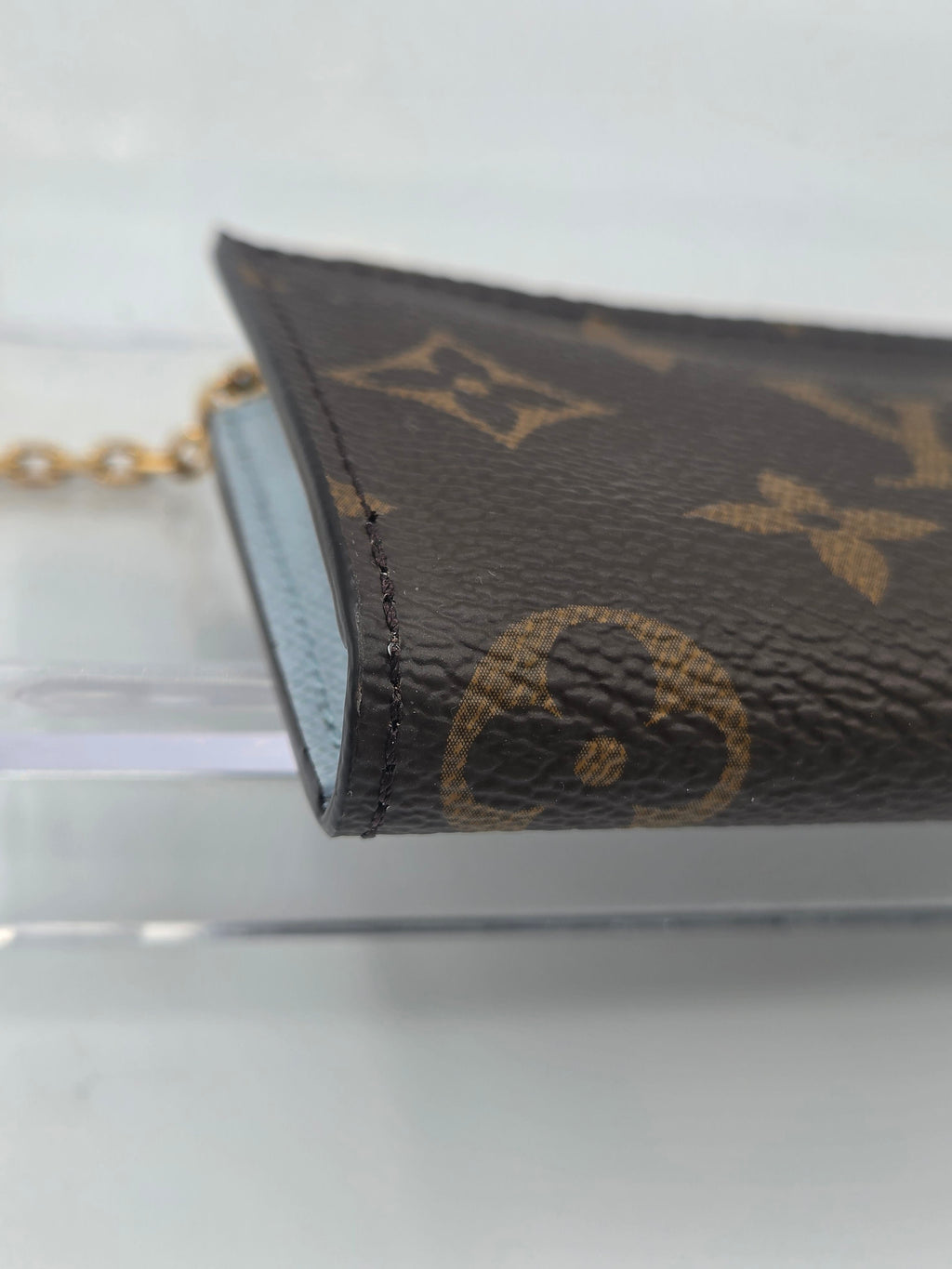 A close-up of a Louis Vuitton monogram canvas keychain or card holder with a gold-tone chain, resting on a clear acrylic stand against a light gray background.