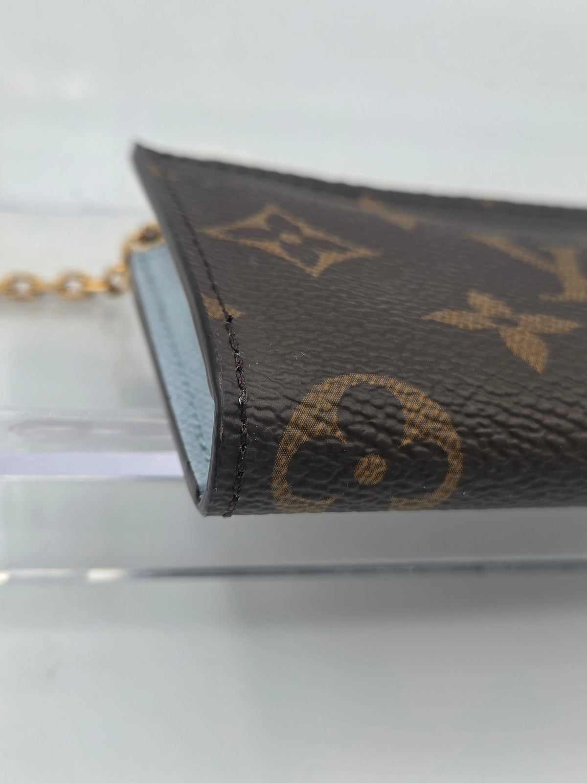 A close-up of a Louis Vuitton monogram canvas keychain or card holder with a gold-tone chain, resting on a clear acrylic stand against a light gray background.