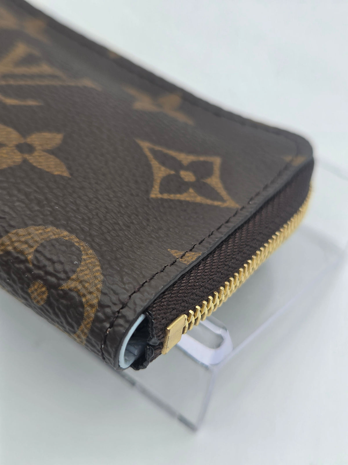 A close-up of a Louis Vuitton monogram zip-around wallet on a clear acrylic stand, showing the classic brown canvas with tan monogram pattern and gold-tone zipper.