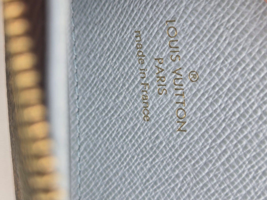 Close-up of a Louis Vuitton product surface with gold 'LOUIS VUITTON made in France' text on a light gray textured canvas, next to a blurred gold zipper edge.
