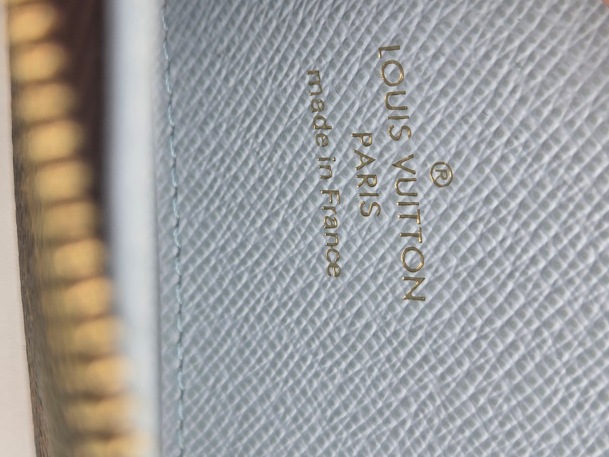 Close-up of a Louis Vuitton product surface with gold 'LOUIS VUITTON made in France' text on a light gray textured canvas, next to a blurred gold zipper edge.