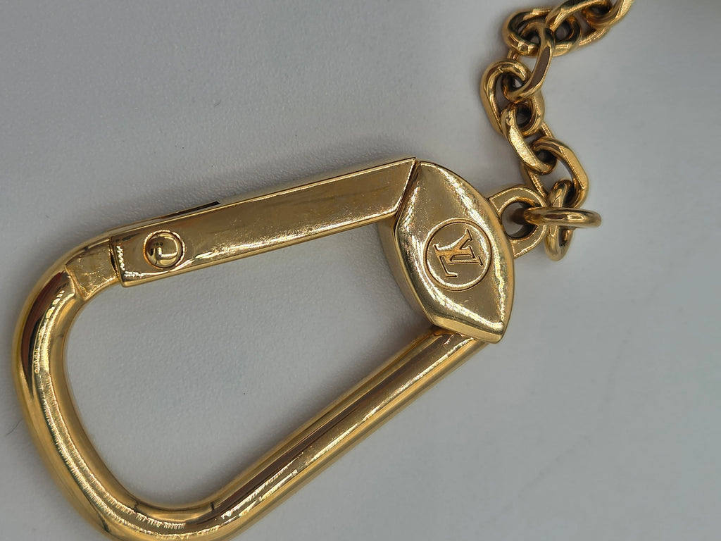 A close-up of a gold-tone metal carabiner with a matching gold chain, featuring a small embossed LV logo on the clasp, set against a plain light gray background.