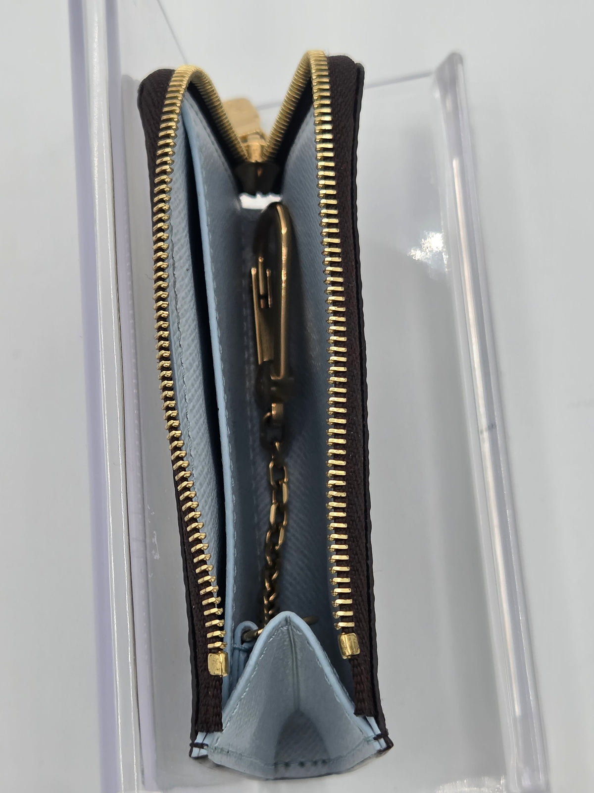 A close-up of a small zippered pouch or wallet with a light blue interior, dark brown exterior, and gold-tone zipper hardware, shown inside a clear plastic display case.