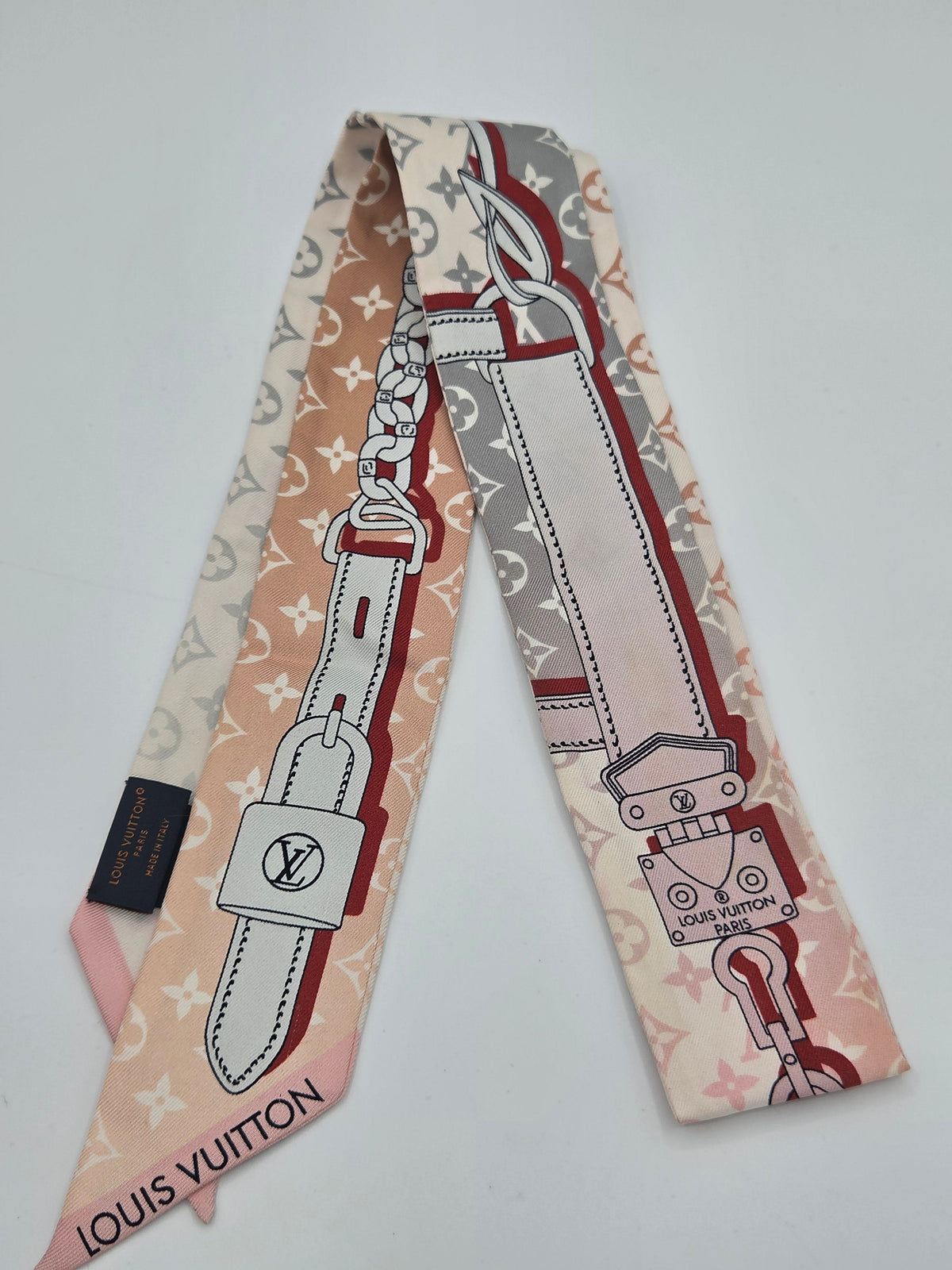 A Louis Vuitton silk scarf featuring a monogram pattern in beige, dusty pink, and gray, with a graphic print of a leather strap and padlock, and the Louis Vuitton name printed on the tip.
