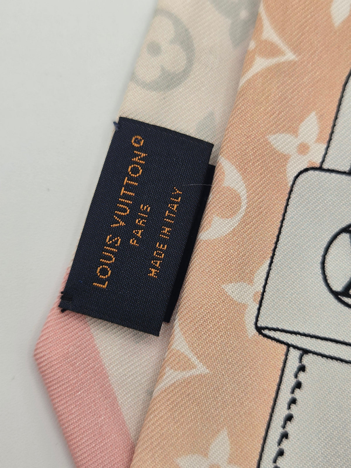 Close-up of a Louis Vuitton scarf showing a navy woven brand label with gold text, alongside pastel pink and peach sections with a monogram-style floral pattern.