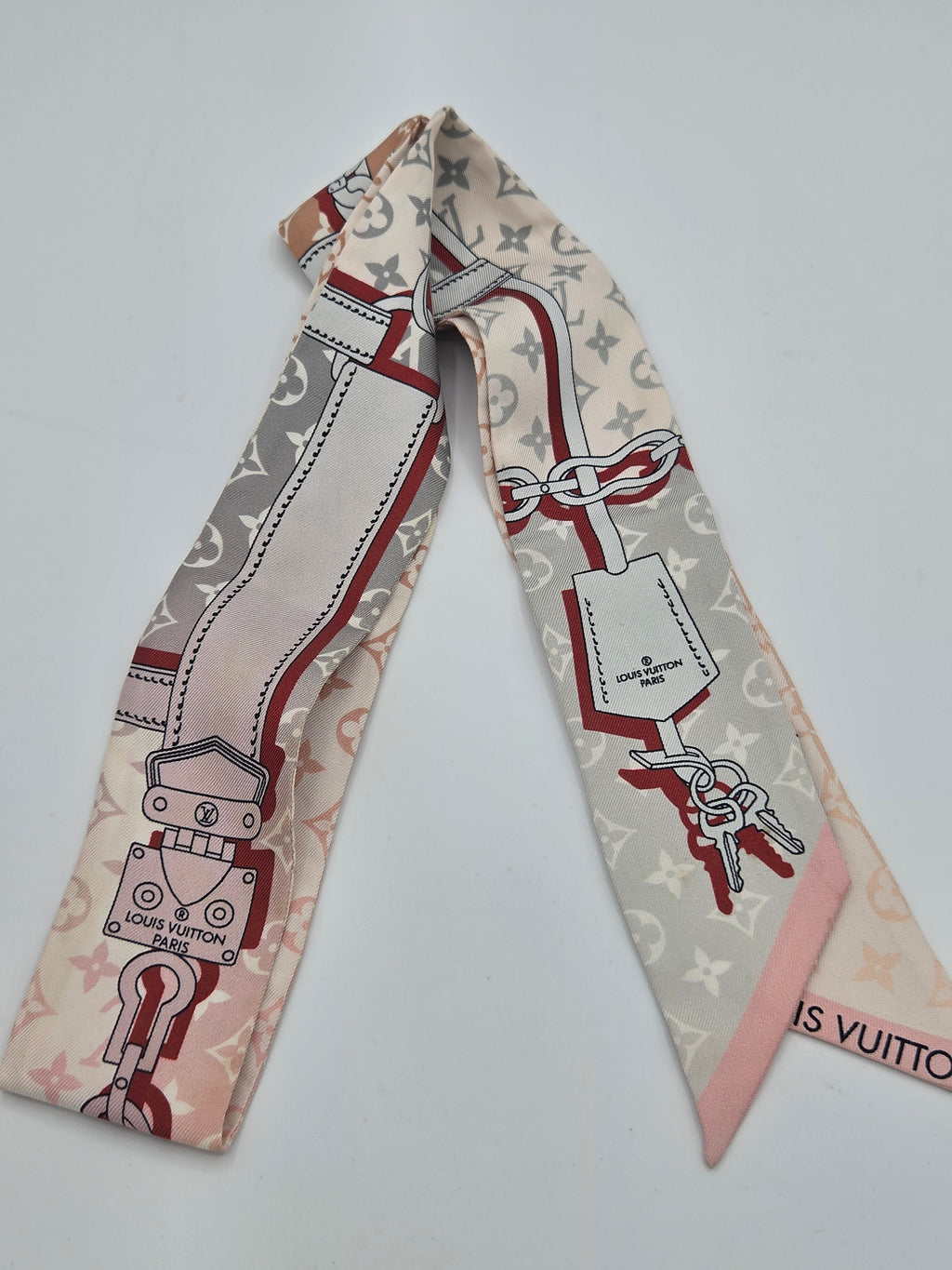 A Louis Vuitton silk scarf featuring the brand's iconic monogram pattern, red and gray border details, and a pink tip, laid flat on a light gray background.