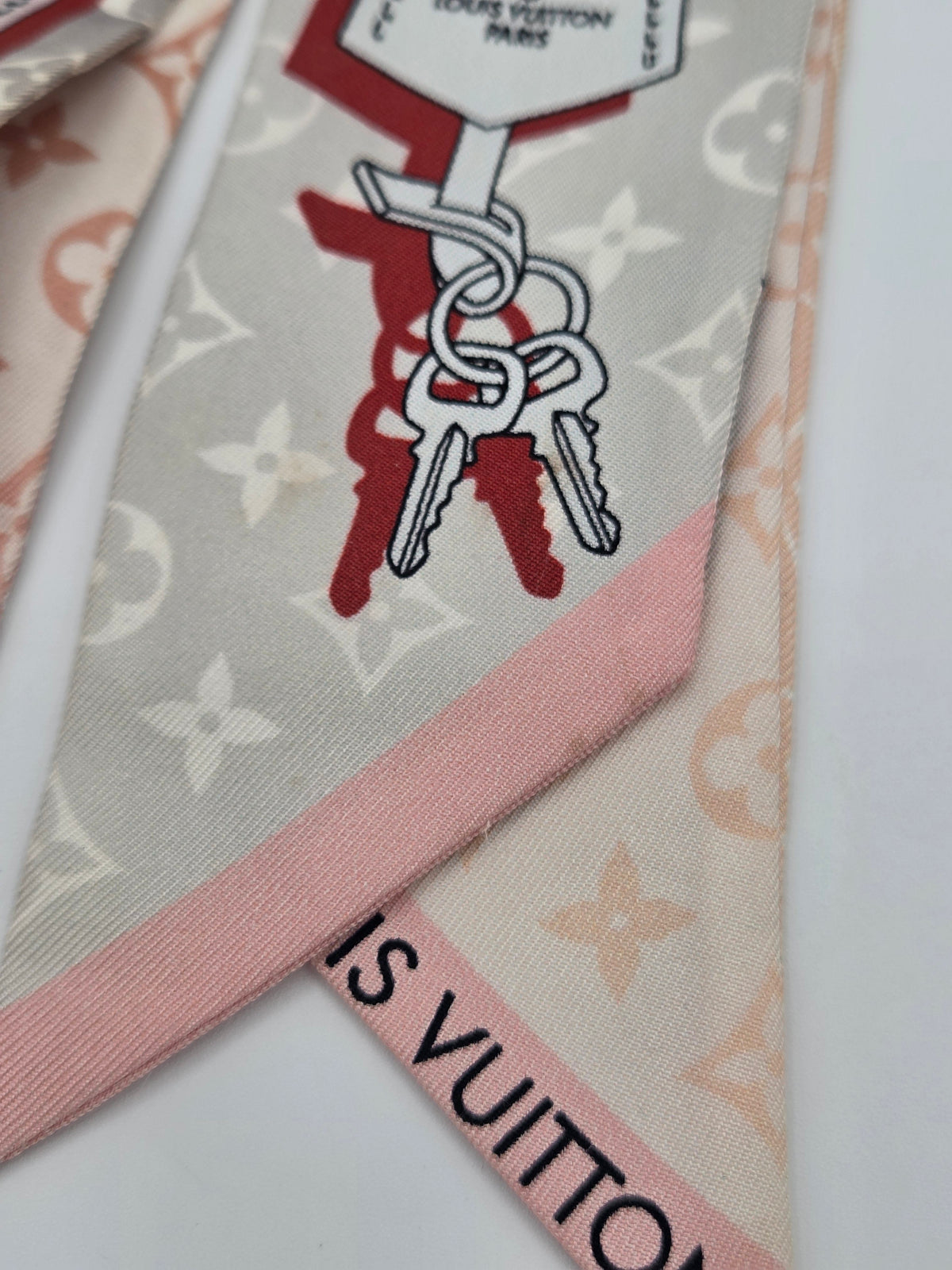 A close-up of a Louis Vuitton silk scarf featuring the brand's classic monogram pattern in beige and peach, a pink border, and a graphic of interlocking keys with a tag.