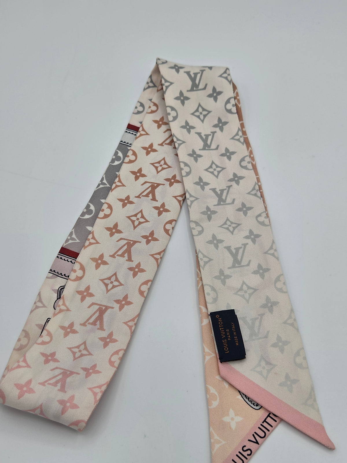 A Louis Vuitton silk scarf with a monogram pattern, featuring a cream base with pink, gray, and brown motifs, and a pink tip with a black brand label.