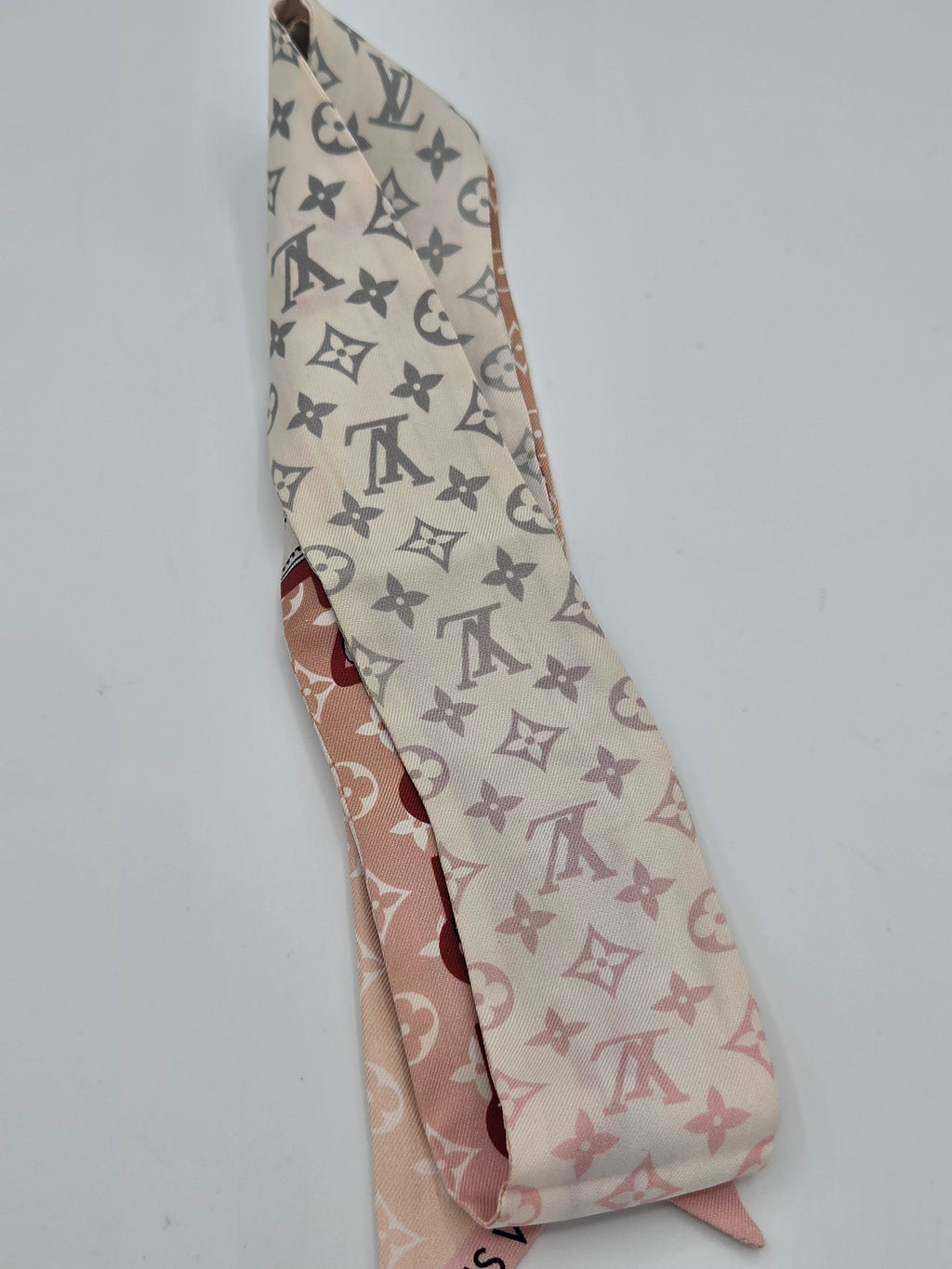 A folded Louis Vuitton silk scarf featuring the classic monogram pattern in gray on a cream background, with a pink reverse side showing the brand name 'LOUIS VUITTON' in red.