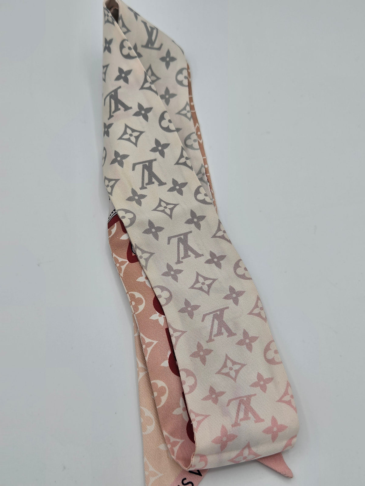 A folded Louis Vuitton silk scarf featuring the classic monogram pattern in gray on a cream background, with a pink reverse side showing the brand name 'LOUIS VUITTON' in red.