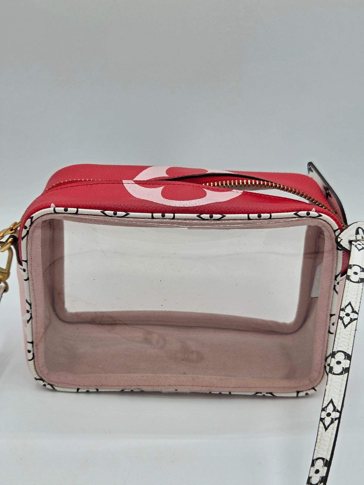 A red camera bag with a clear front window, a pink interior lining, and a white strap featuring a black monogram pattern. A large white logo is printed on the red top flap.