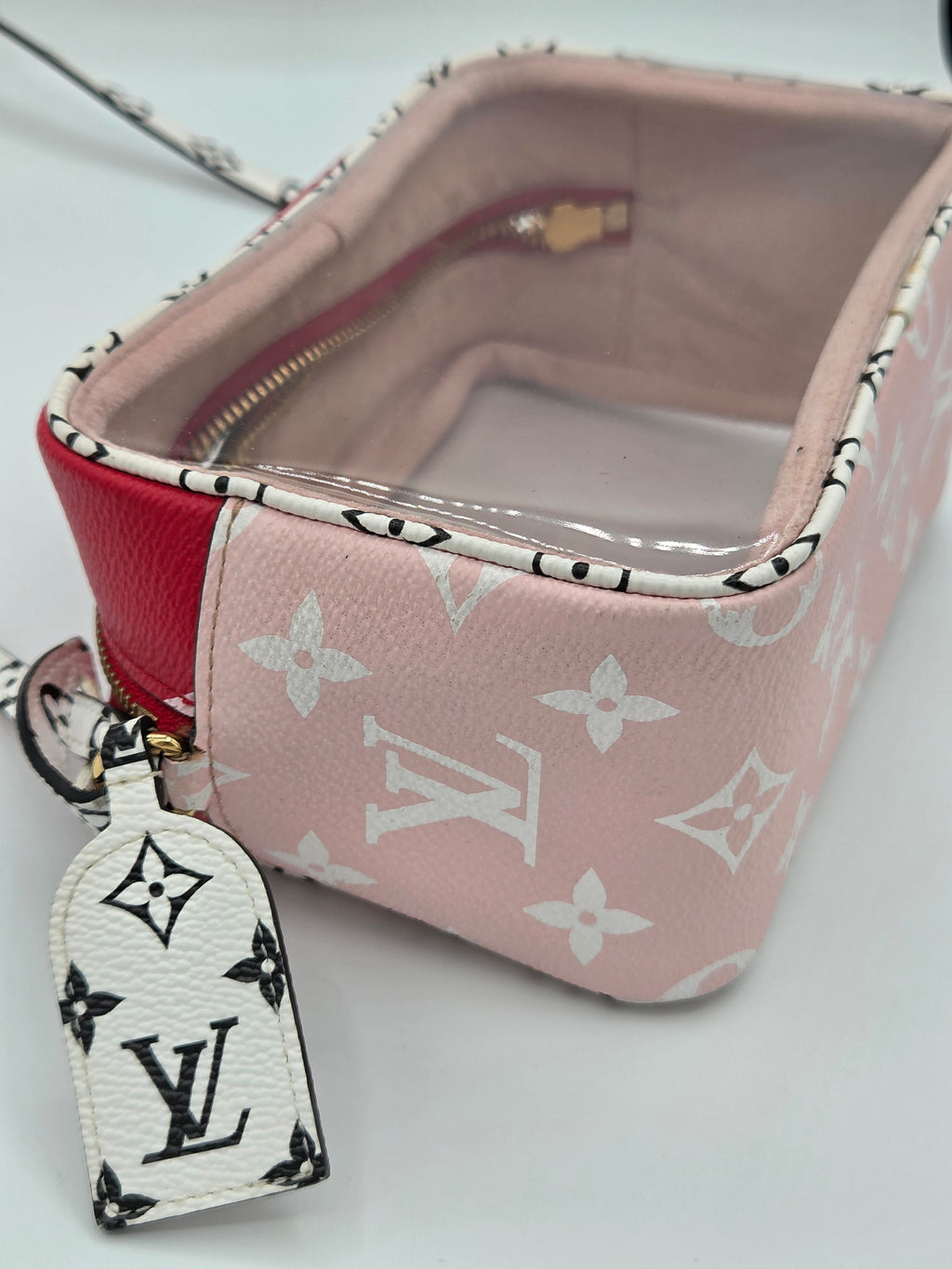 A Louis Vuitton monogram toiletry bag in light pink and white, featuring a red leather side panel, a clear zippered top, and a white monogram luggage tag hanging from the zipper.