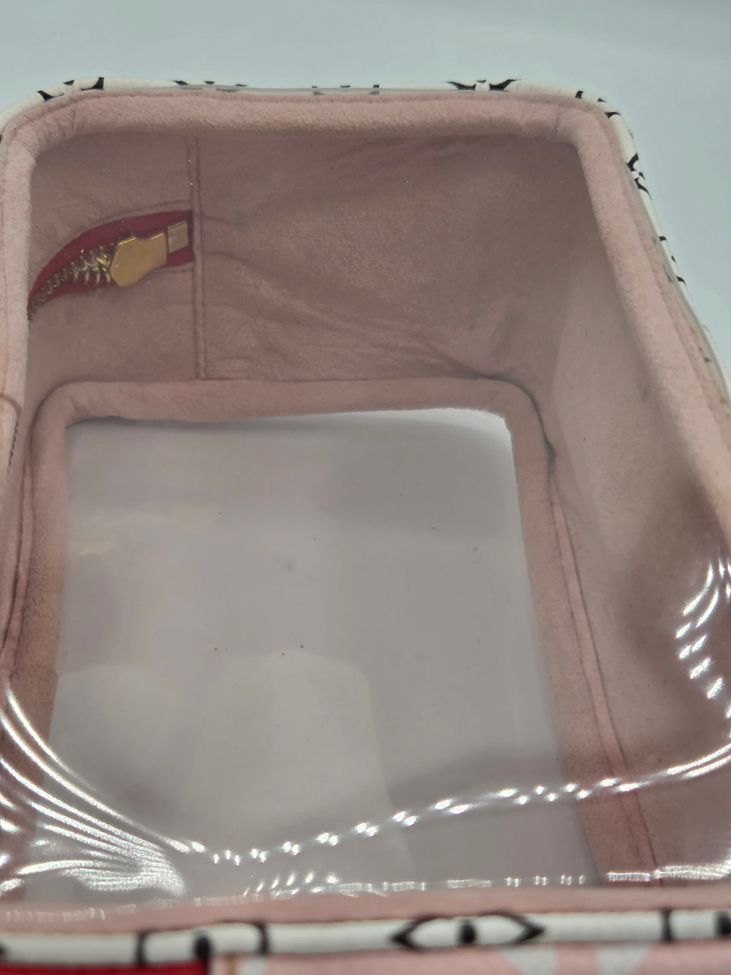 A square pink fabric storage bin with a clear plastic window on the front, showing a dusty white surface inside. The bin has a patterned fabric trim at the top and a small red and gold tag inside.
