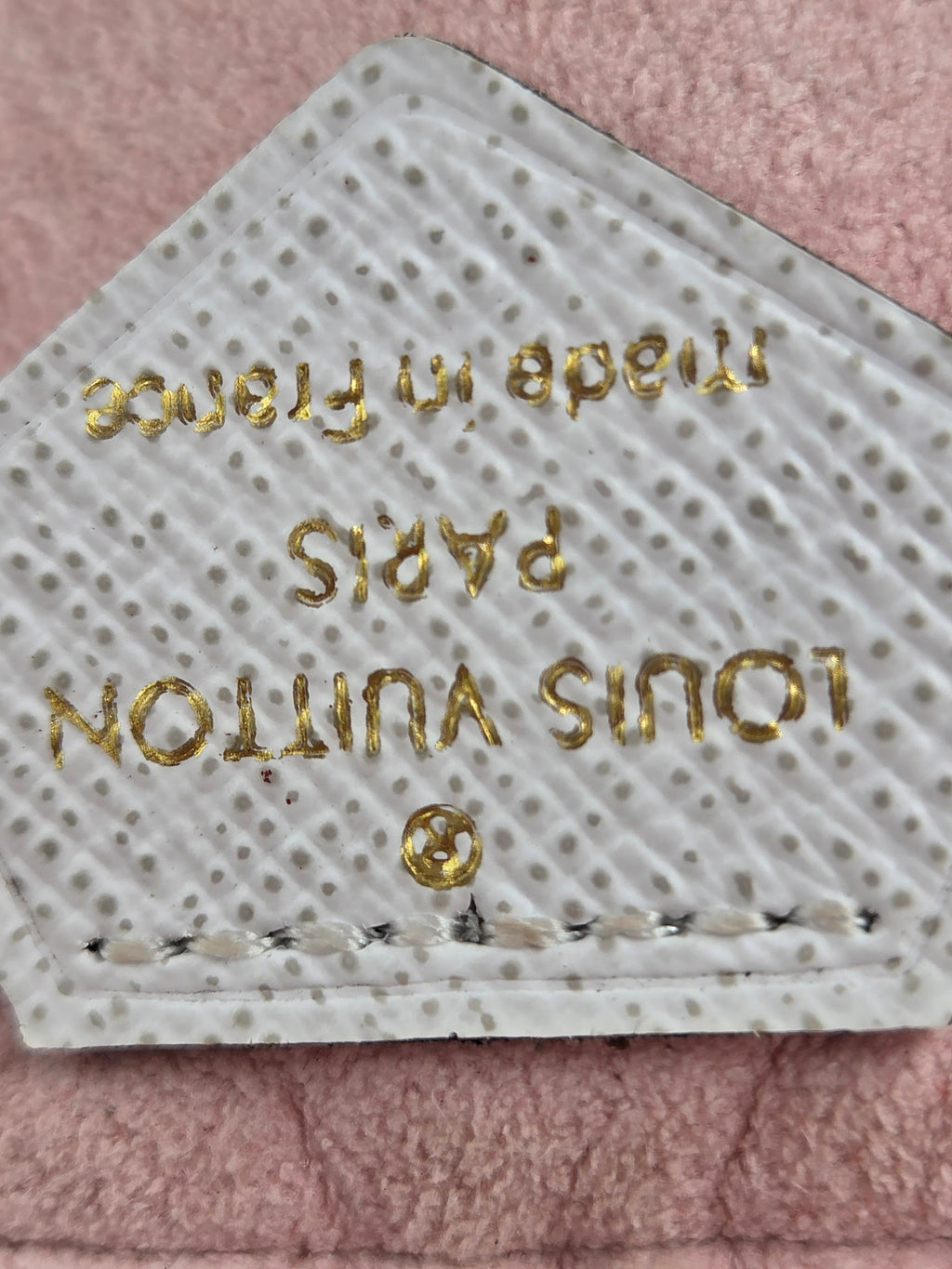 A close-up of a Louis Vuitton leather patch with gold foil branding and 'Made in France' text, placed on a pink textured fabric background.