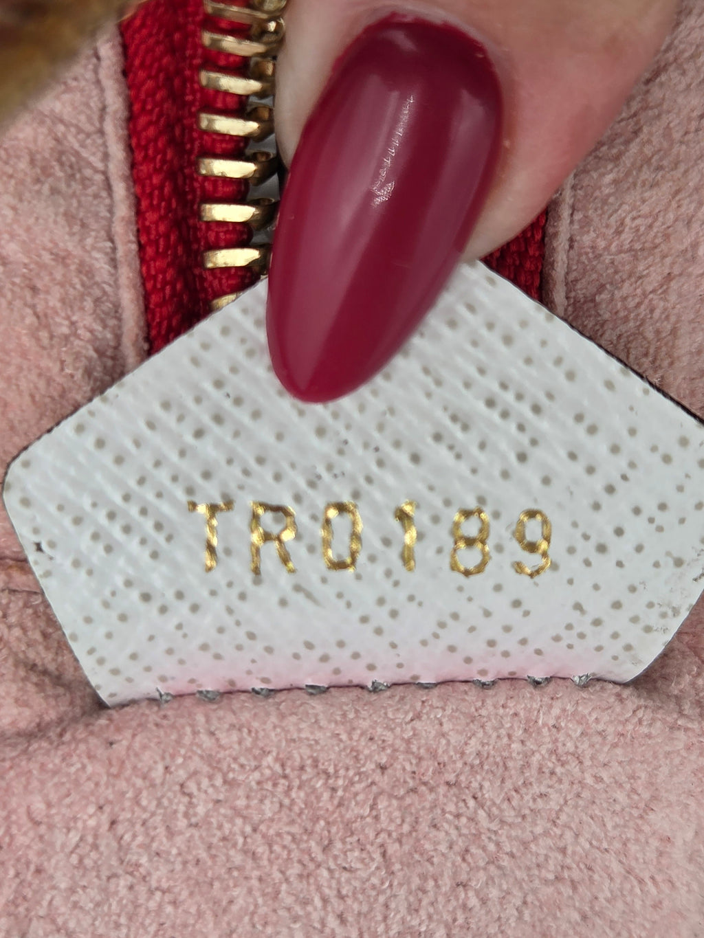 Close-up of a hand with dark red manicured nails pressing down on a white textured patch with gold-foiled lettering that reads 'TRO189', positioned over a pink fabric and red zipper.