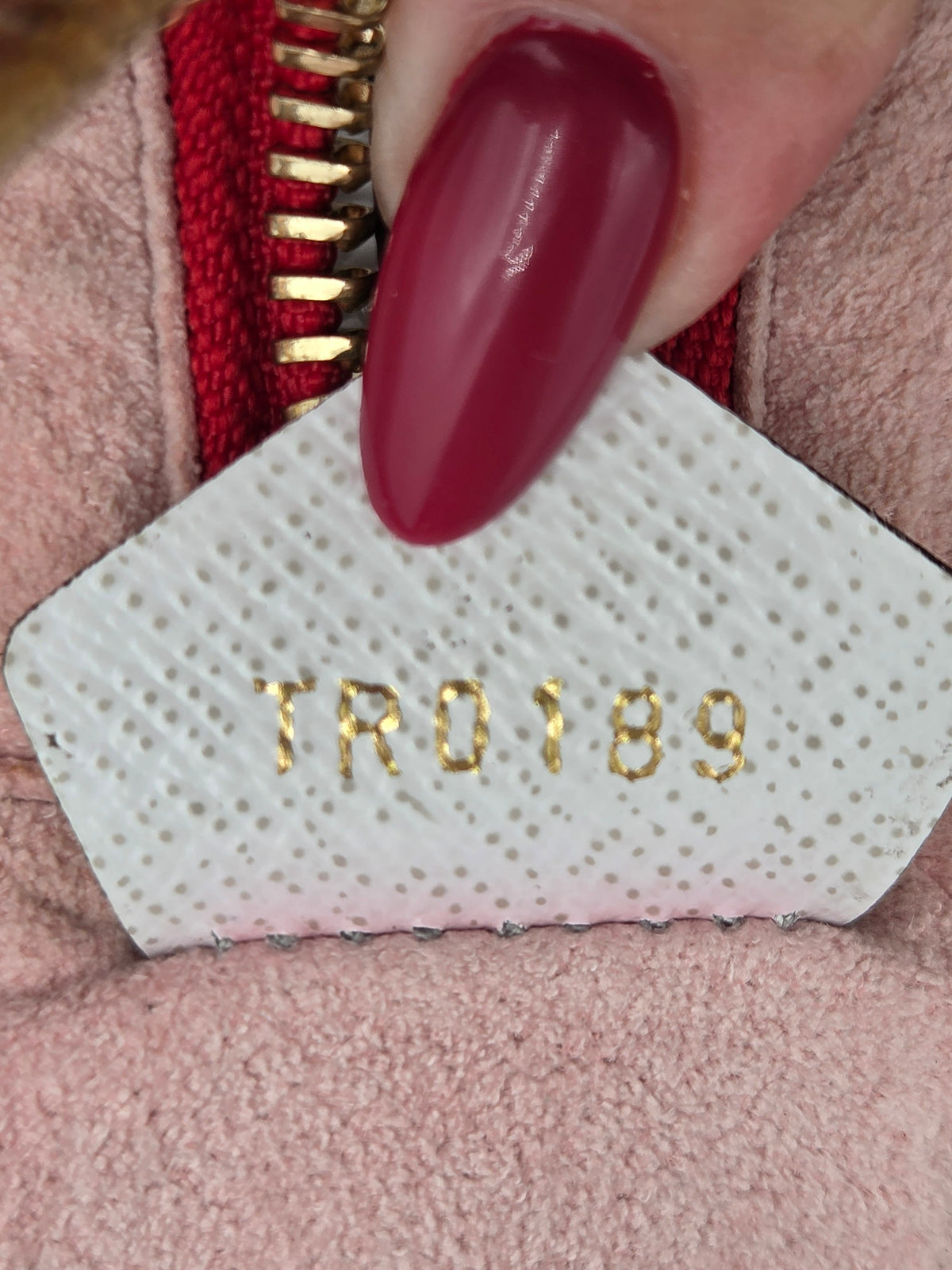 Close-up of a hand with dark red manicured nails pressing down on a white textured patch with gold-foiled lettering that reads 'TRO189', positioned over a pink fabric and red zipper.
