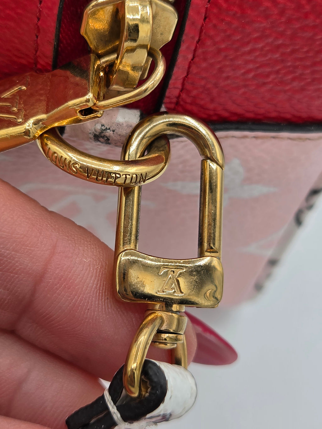 Close-up of a Louis Vuitton bag hardware clasp held by a hand, showing the gold-tone padlock-shaped clasp engraved with 'LOUIS VUITTON' and an LV logo, attached to a red textured leather strap.
