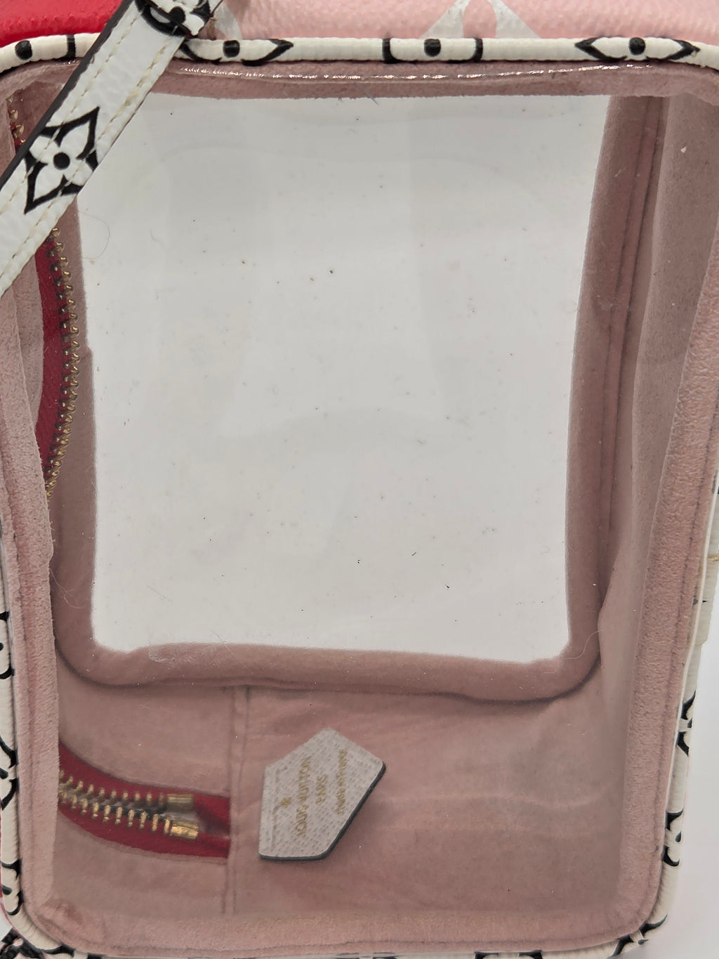 A close-up of a pink cosmetic pouch with a clear plastic window, a patterned fabric trim, a red zipper pocket, and a small white brand tag.