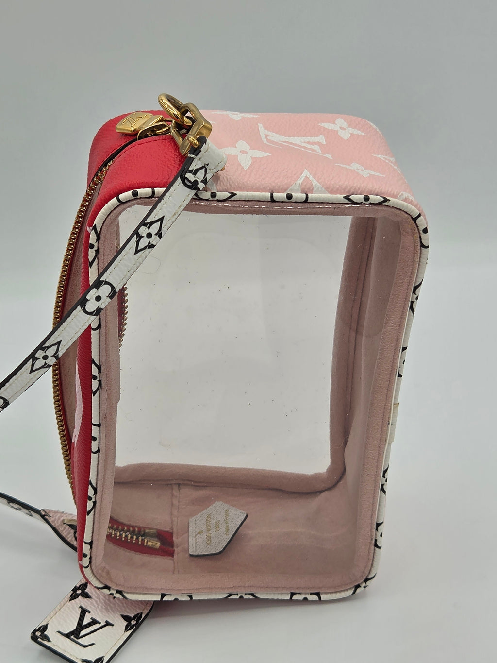 A Louis Vuitton clear plastic pouch with a pink monogram canvas exterior, a red zippered pocket, and a white monogram shoulder strap, shown on a neutral gray background.