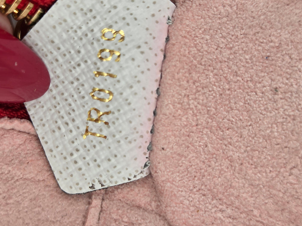 A close-up of a light pink textured fabric item, likely a shoe or bag, showing a white perforated patch with gold foil stamping that reads 'TRO198'. A dark pink fingernail is visible on the left edge.