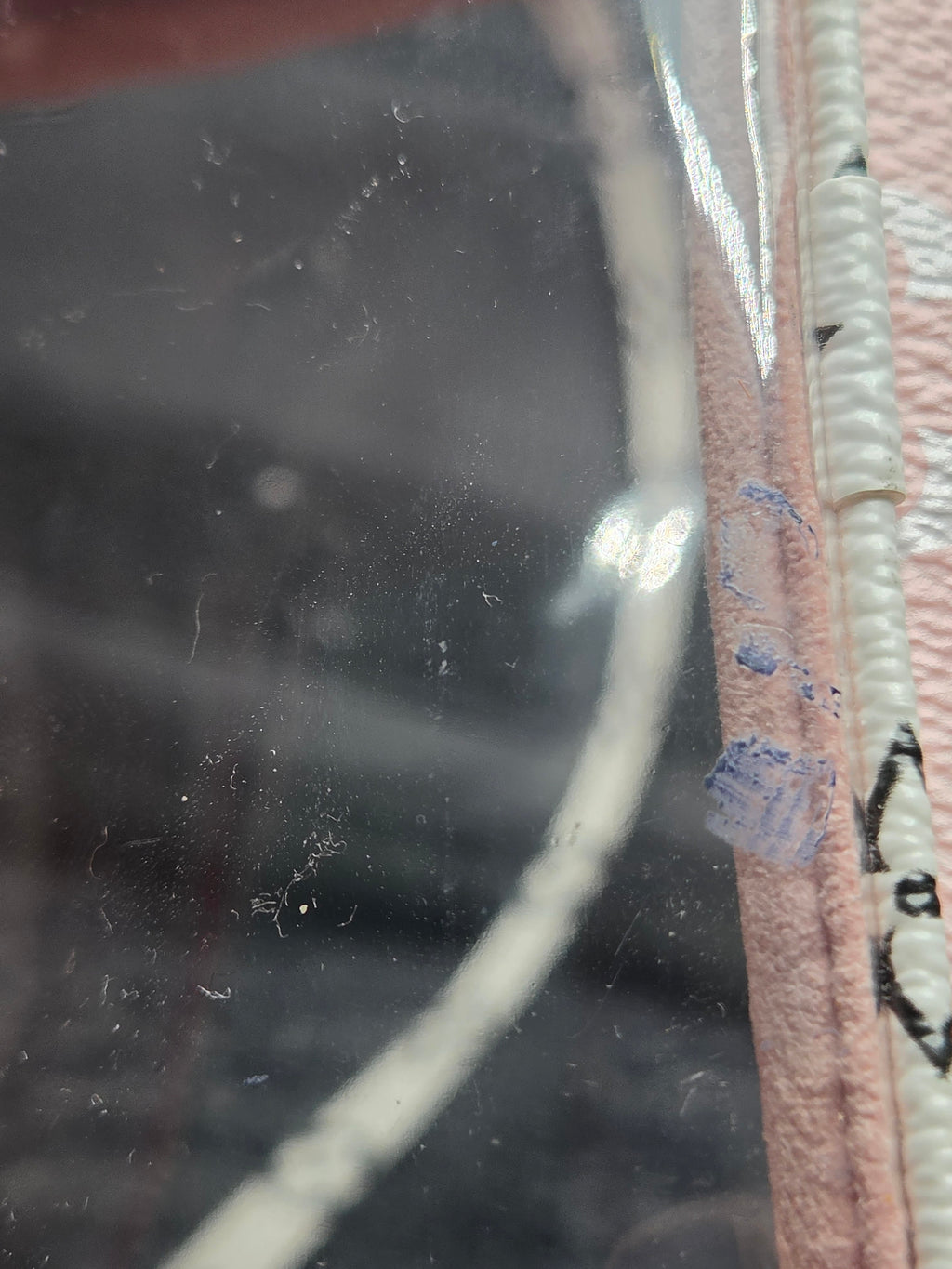 Close-up of a pink faux-leather accessory with a clear plastic window and white trim, showing visible dust, scratches, and blue paint marks on the clear surface.