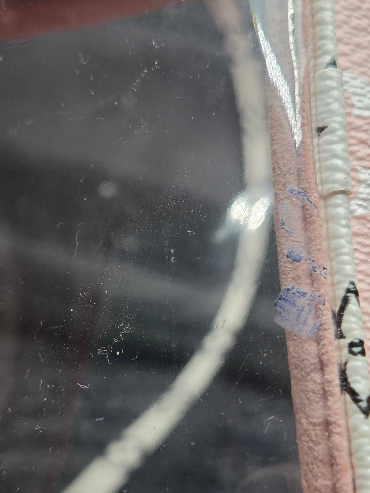 Close-up of a pink faux-leather accessory with a clear plastic window and white trim, showing visible dust, scratches, and blue paint marks on the clear surface.