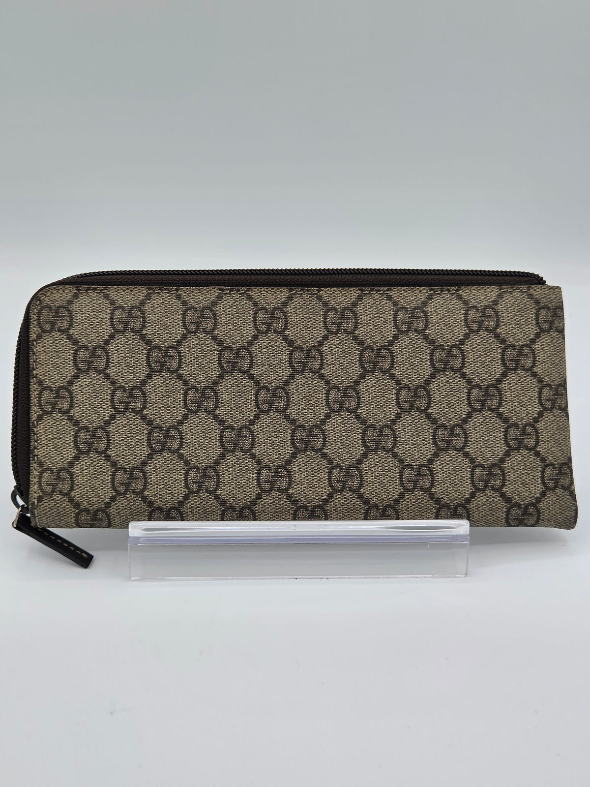 A Gucci zip-around pouch in the brand's signature GG Supreme canvas, featuring a beige and brown monogram pattern, displayed on a clear acrylic stand against a light gray background.