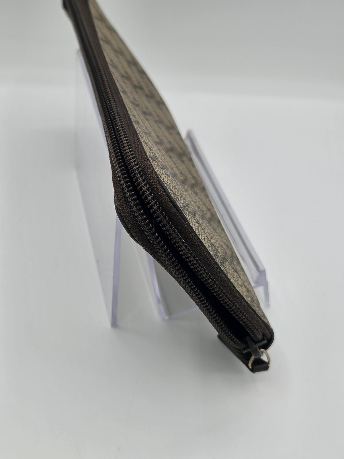 A close-up of a zippered pouch with a brown and beige monogram pattern, displayed on a clear acrylic stand against a white background.