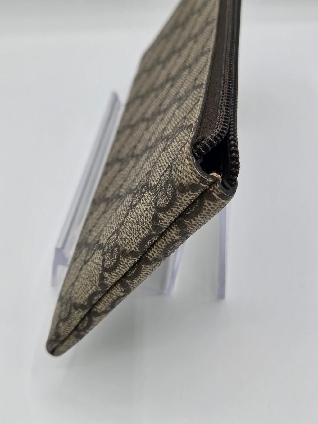 A close-up of a beige and brown monogram canvas zippered pouch standing on a clear acrylic display stand against a white background.