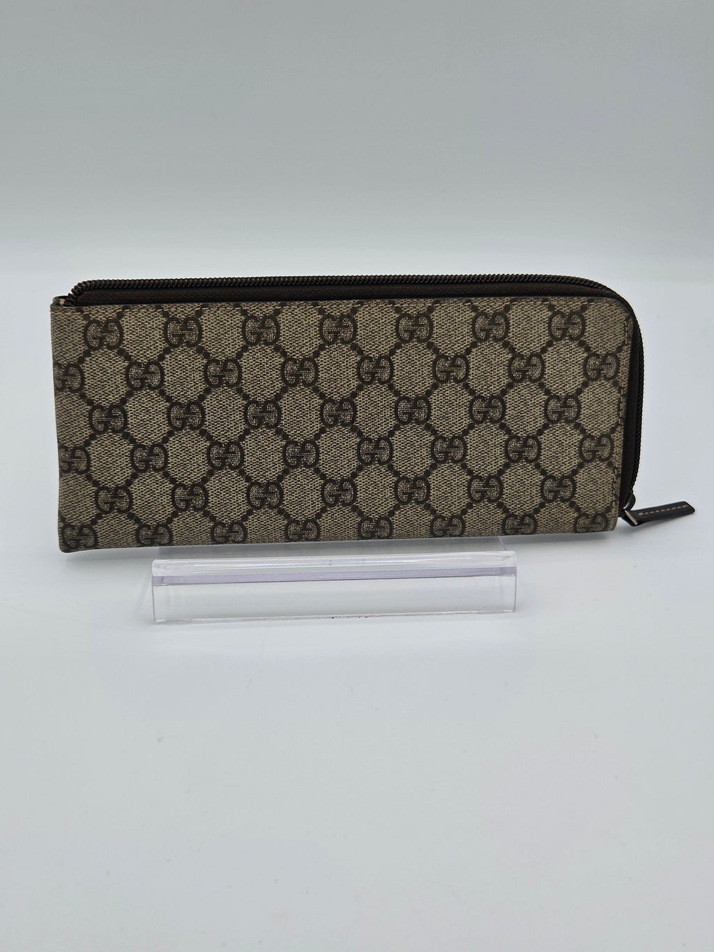 A Gucci zip-around pouch in the brand's signature GG monogram canvas, displayed on a clear acrylic stand against a neutral light gray background.